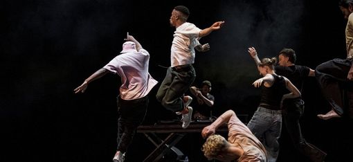 Several dancers in dynamic poses on a dark stage, two jumping in the air, one kneeling bent forward