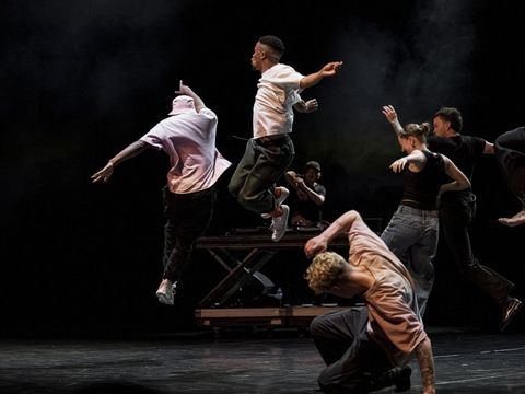 Several dancers in dynamic poses on a dark stage, two jumping in the air, one kneeling bent forward