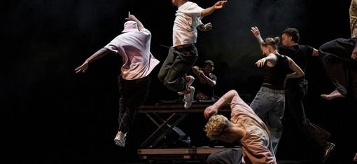 Several dancers in dynamic poses on a dark stage, two jumping in the air, one kneeling bent forward