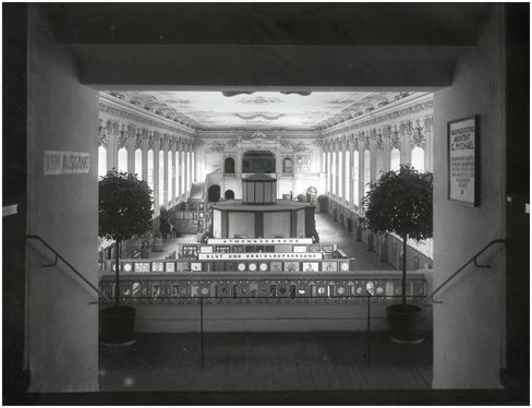 Blick durch einen Torbogen auf eine große, prunkvolle Messehalle mit hohen Fenstern, dekorativer Decke und mehreren Ausstellungsständen, flankiert von zwei großen Pflanzen.