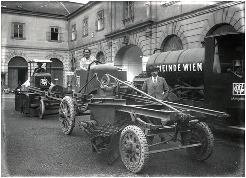 Historisches Schwarz-weiß-Foto mit zwei landwirtschaftlichen Maschinen und einem Tankwagen mit der Aufschrift 'GEMEINDE WIEN' in einem Innenhof mit Arkaden.