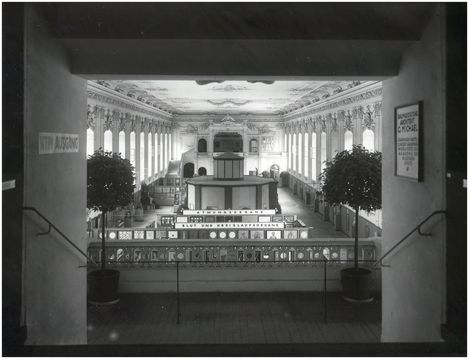 Blick durch einen Torbogen auf eine große, prunkvolle Messehalle mit hohen Fenstern, dekorativer Decke und mehreren Ausstellungsständen, flankiert von zwei großen Pflanzen.