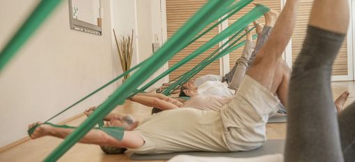 Mehrere Personen liegen auf Matten und dehnen ihre Beine mit grünen Widerstandsbändern in einem hellen Raum