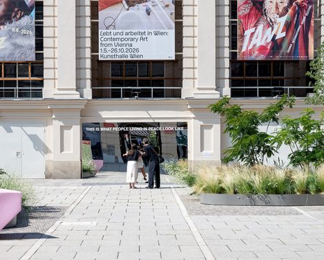 Großer Spiegel mit der Aufschrift 'THIS IS WHAT PEOPLE LIVING IN PEACE LOOK LIKE' vor einem historischen Gebäude mit zwei großen Bannern und mehreren Personen davor