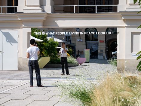 Large mirror with the text 'THIS IS WHAT PEOPLE LIVING IN PEACE LOOK LIKE' reflecting a person wearing black pants and a white t-shirt, outdoors in front of a light-colored building with plants and paving stones.