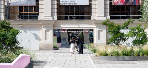 Großer Spiegel mit der Aufschrift 'THIS IS WHAT PEOPLE LIVING IN PEACE LOOK LIKE' vor einem historischen Gebäude mit zwei großen Bannern und mehreren Personen davor