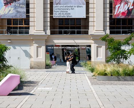 Two people are hugging in front of a historic building with three large banners, in front of a mirror bearing the words ‘THIS IS WHAT PEOPLE LIVING IN PEACE LOOK LIKE’