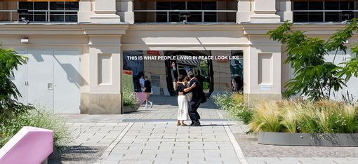 Zwei Personen umarmen sich vor einem historischen Gebäude mit drei großen Bannern vor einem Spiegel mit dem Schriftzug 'THIS IS WHAT PEOPLE LIVING IN PEACE LOOK LIKE'