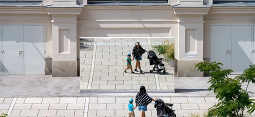 Großer Spiegel mit der Aufschrift 'THIS IS WHAT PEOPLE LIVING IN PEACE LOOK LIKE', der eine Frau mit Kinderwagen und ein Kind auf einem gepflasterten Platz reflektiert.