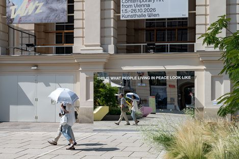 Spiegel mit der Aufschrift 'THIS IS WHAT PEOPLE LIVING IN PEACE LOOK LIKE' vor einem historischen Gebäude mit Menschen, die daran vorbeigehen, und einer weißen Sonnenschirmattrappe.