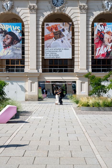 Zwei Personen umarmen sich vor einem historischen Gebäude mit drei großen Bannern vor einem Spiegel mit dem Schriftzug 'THIS IS WHAT PEOPLE LIVING IN PEACE LOOK LIKE'