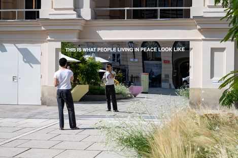 Großer Spiegel mit der Aufschrift 'THIS IS WHAT PEOPLE LIVING IN PEACE LOOK LIKE', der eine Person in schwarzer Hose und weißem T-Shirt reflektiert, im Außenbereich vor einem hellen Gebäude mit Pflanzen und Pflastersteinen.