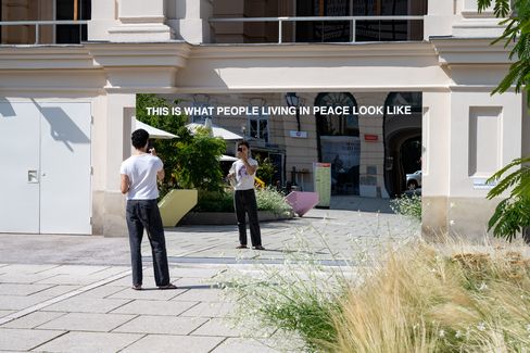 Großer Spiegel mit der Aufschrift 'THIS IS WHAT PEOPLE LIVING IN PEACE LOOK LIKE', der eine Person in schwarzer Hose und weißem T-Shirt reflektiert, im Außenbereich vor einem hellen Gebäude mit Pflanzen und Pflastersteinen.
