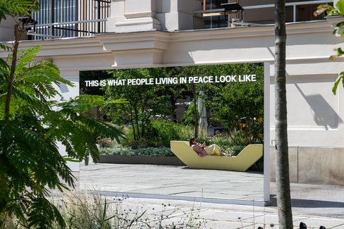 Großer rechteckiger Spiegel mit der Aufschrift 'THIS IS WHAT PEOPLE LIVING IN PEACE LOOK LIKE' in Großbuchstaben, der eine Person zeigt, die auf einer modernen Bank in einem urbanen Gartenbereich liegt.