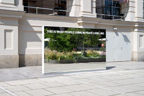 Großer rechteckiger Spiegel mit der weißen Aufschrift 'THIS IS WHAT PEOPLE LIVING IN PEACE LOOK LIKE' vor einem historischen Gebäude auf einem gepflasterten Platz