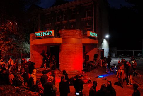 Crowd in front of a rounded brick building with illuminated signs and red lighting at night.