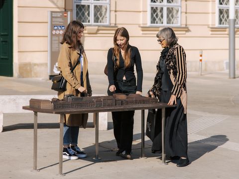 Drei Personen stehen um einen Tisch mit einem Modell des MuseumsQuartier Wien auf dem MQ Vorplatz mit einer hellen Fassade.