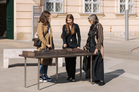 Drei Personen stehen um einen Tisch mit einem Modell des MuseumsQuartier Wien auf dem MQ Vorplatz mit einer hellen Fassade.