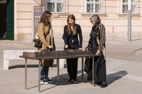 Drei Personen stehen um einen Tisch mit einem Modell des MuseumsQuartier Wien auf dem MQ Vorplatz mit einer hellen Fassade.