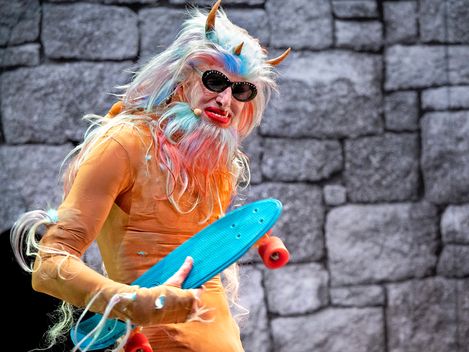 Ein Monster mit drei Hörnern, Sonnenbrille, verlängertem Eckzahn und einem Skateboard in der Hand schaut direkt in die Kamera.