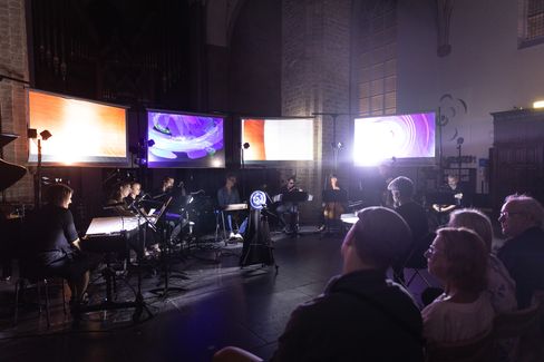Musicians seated in a dark room in front of four large screens showing abstract colorful projections with audience in the foreground.