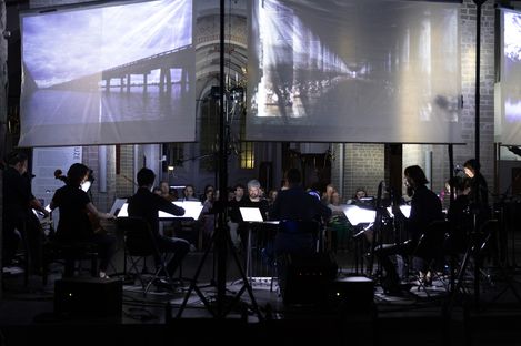 Musicians sit in darkness with illuminated music stands in front of large screens showing bridge projections.