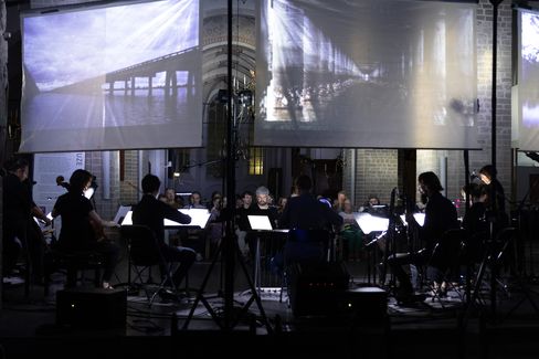 Musicians sit in darkness with illuminated music stands in front of large screens showing bridge projections.