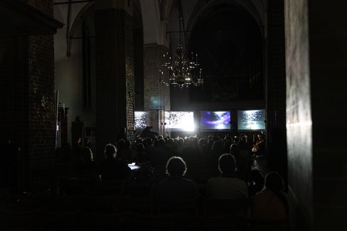 Dark interior of a church with an audience facing three illuminated screens and a chandelier hanging from the ceiling.