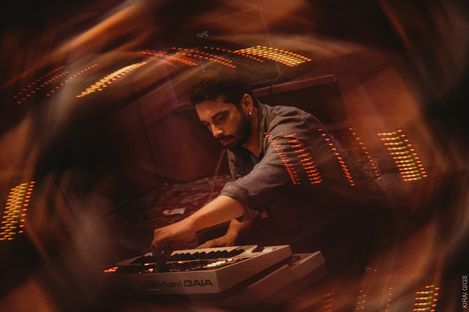 Person with dark hair operating a Roland GAIA synthesizer keyboard in a room with blurred orange lights in the foreground.