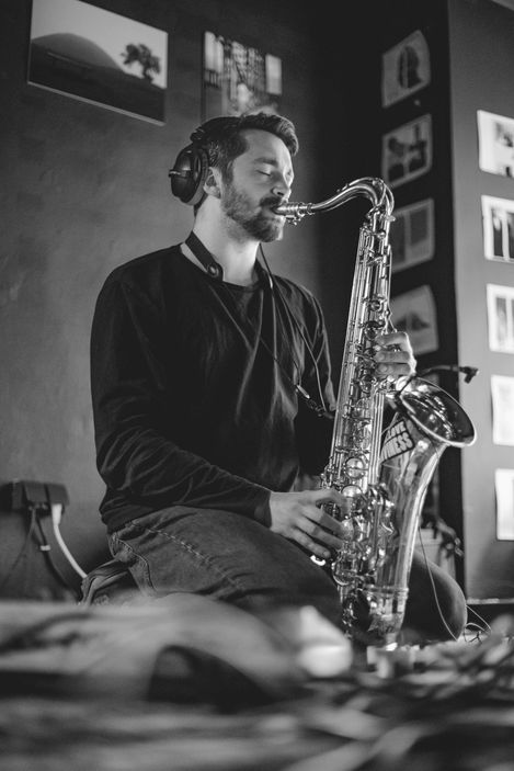 Black-and-white photo of a seated man wearing headphones playing a saxophone in front of a wall with multiple pictures.