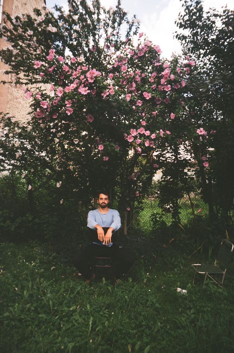 Bearded man sitting on a chair under a flowering tree in a garden.