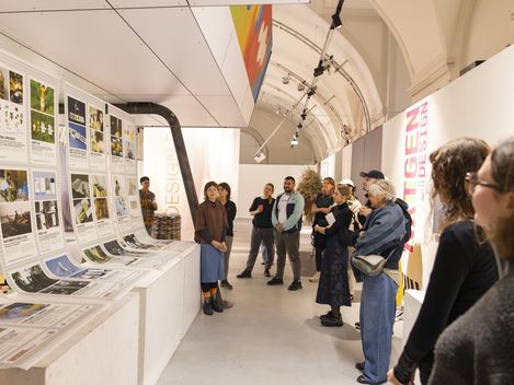 Gruppe von Menschen in einem Ausstellungsraum mit hohen gewölbten Decken, die an einer Wand mit ausgedruckten Plakaten und Bildern stehen