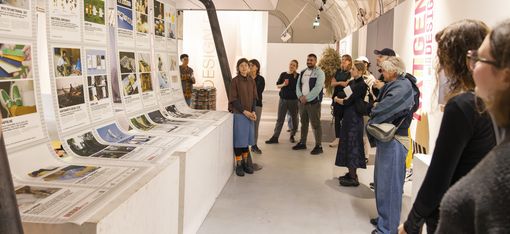 Gruppe von Menschen in einem Ausstellungsraum mit hohen gewölbten Decken, die an einer Wand mit ausgedruckten Plakaten und Bildern stehen