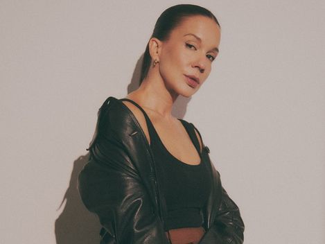 Woman with pulled-back hair wearing black top, black leather jacket, and brown pants sitting against a light wall
