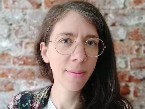 Person with long dark hair wearing a white top and a black floral cover in front of a brick wall.