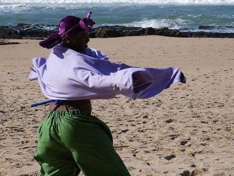 Person wearing a purple headscarf and loose purple top dancing on a sandy beach wearing green pants