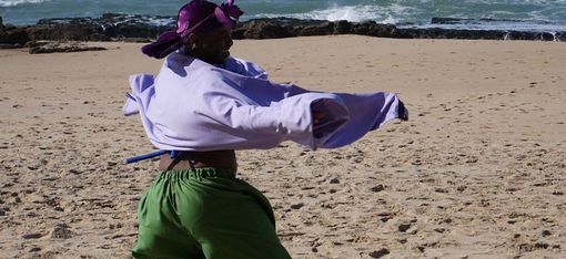 Person wearing a purple headscarf and loose purple top dancing on a sandy beach wearing green pants