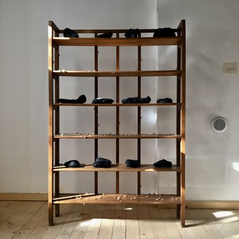 Wooden shelf with several black irregularly shaped objects and small light-colored spheres on the shelves against a white wall and wooden floor.