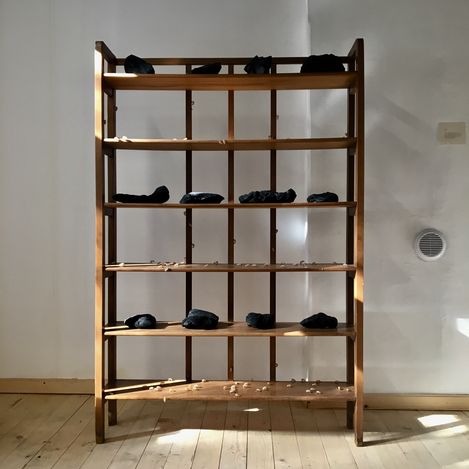 Wooden shelf with several black irregularly shaped objects and small light-colored spheres on the shelves against a white wall and wooden floor.