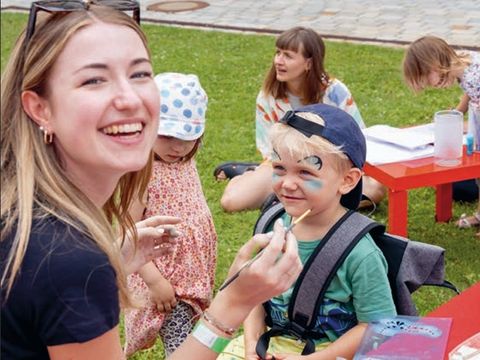 Woman painting a boy's face outdoors with a brush while other children sit at red tables.