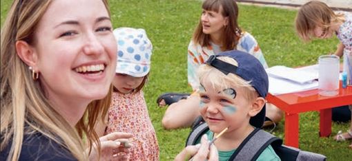 Frau malt einem Jungen im Freien mit einem Pinsel das Gesicht während weitere Kinder an roten Tischen sitzen.