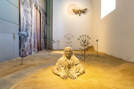 Sculpture of a seated person with a covered head and hands in sand, surrounded by stylized metal plants and a large butterfly shape on the wall.