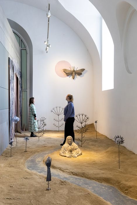 Two people stand in a sand-covered room with sculptures of plants, a seated figure, and a large insect sculpture on the wall.
