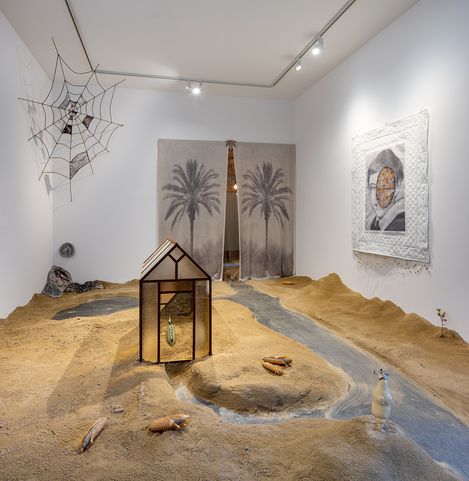 Art installation with a model house, sandy landscape, artificial stream, several bottles, a spiderweb sculpture, and two wall artworks featuring palm trees and geometric pattern.