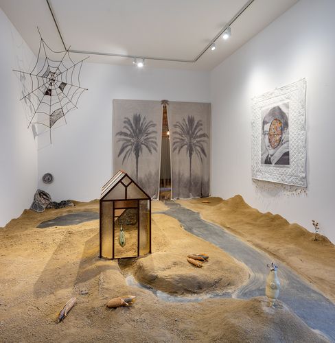 Art installation with a model house, sandy landscape, artificial stream, several bottles, a spiderweb sculpture, and two wall artworks featuring palm trees and geometric pattern.