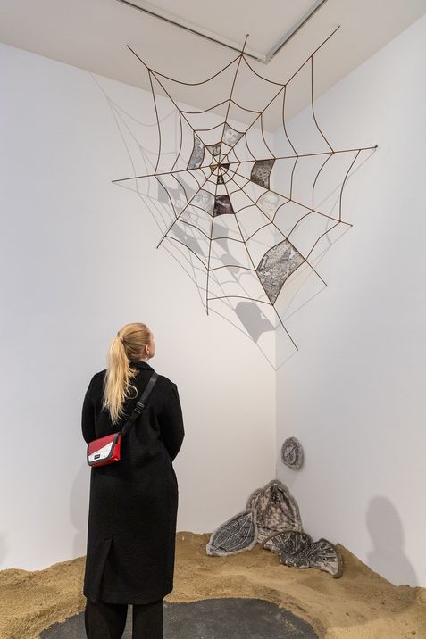 Woman with blond ponytail and red shoulder bag stands in front of a large spiderweb sculpture on a white wall with sand-covered floor and several round objects
