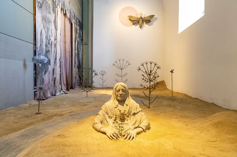 Sculpture of a seated person with a covered head and hands in sand, surrounded by stylized metal plants and a large butterfly shape on the wall.