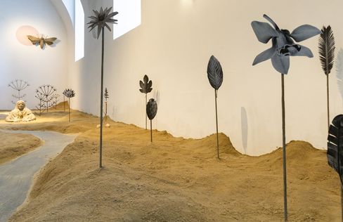 Multiple metal plant shapes stand on sandy ground in an exhibition space.