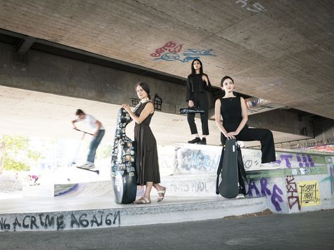 Three women dressed in black with guitar cases stand and sit on a graffiti-covered concrete ramp under a bridge with a skateboarder in motion in the background