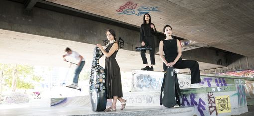 Drei Frauen in schwarzer Kleidung mit Gitarrentaschen stehen und sitzen auf einer mit Graffiti bedeckten Betonrampe unter einer Brücke, im Hintergrund ein Skateboarder in Bewegung